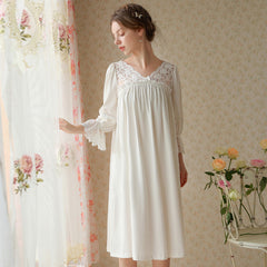 Woman wearing a white lace nightgown in a softly lit room with floral curtains and flowers.